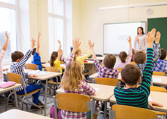 young students sitting at desks in a classroom and raising their hands